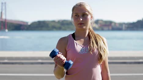 Sporty-young-woman-exercising-with-dumbbells-on-riverside.